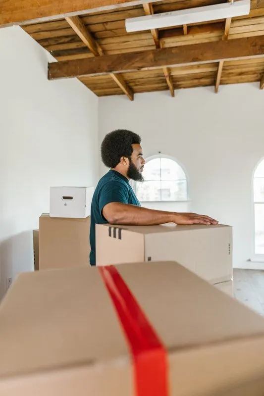 Mover in teal shirt standing among moving boxes in an empty room (stock photo)
